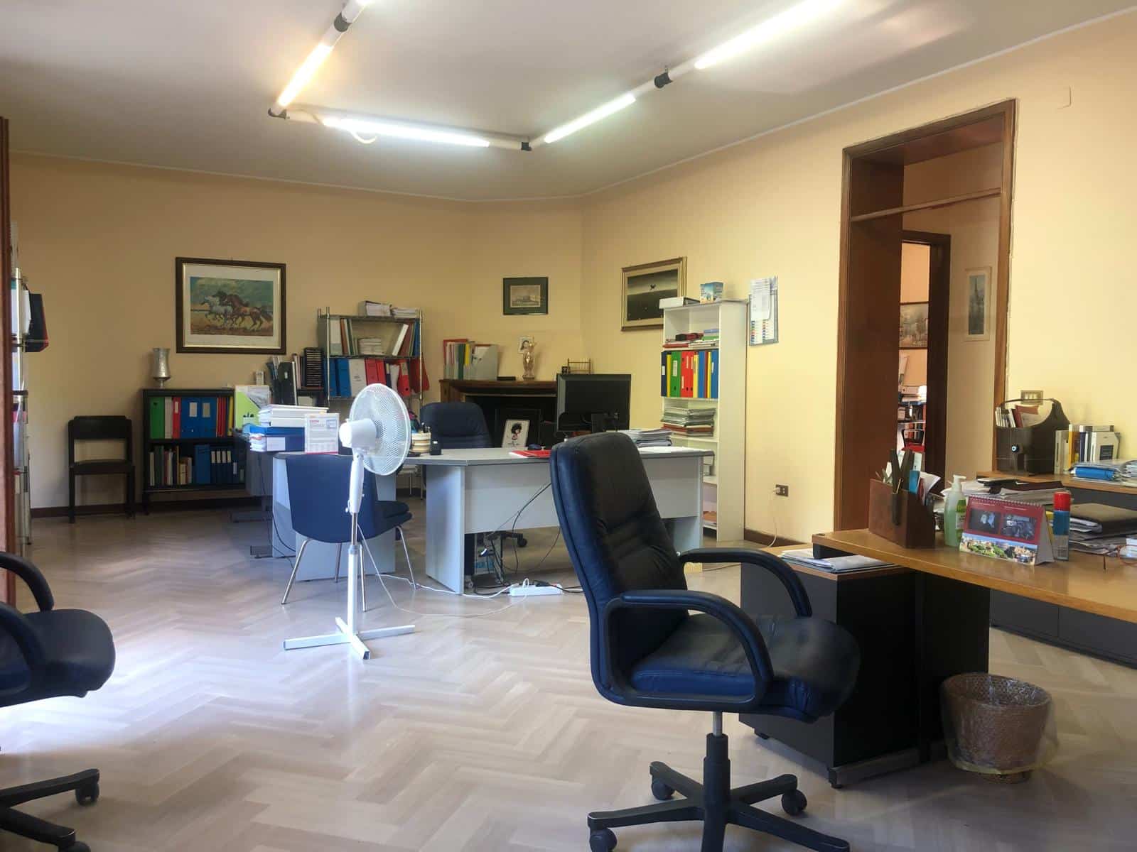 Spazio ufficio con scrivanie, sedie e scaffali pieni di documenti e libri, ambiente di lavoro ordinato e funzionale, lampade a soffitto, decorazioni alle pareti, ideale per professionisti e studi di lavoro.