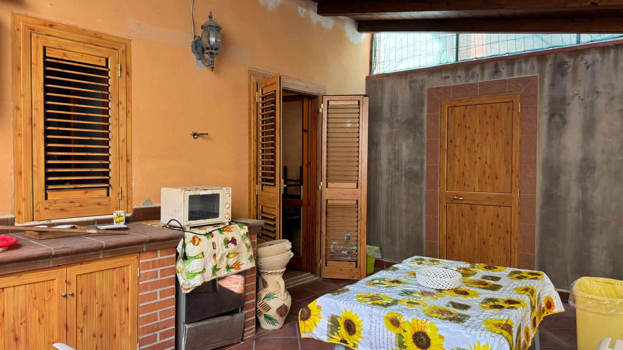 Finestra in legno con persiana scorrevole e cucina all'aperto in stile rustico con tavolo da pranzo decorato con tovaglia a girasole, ambiente esterno accogliente e ben curato.