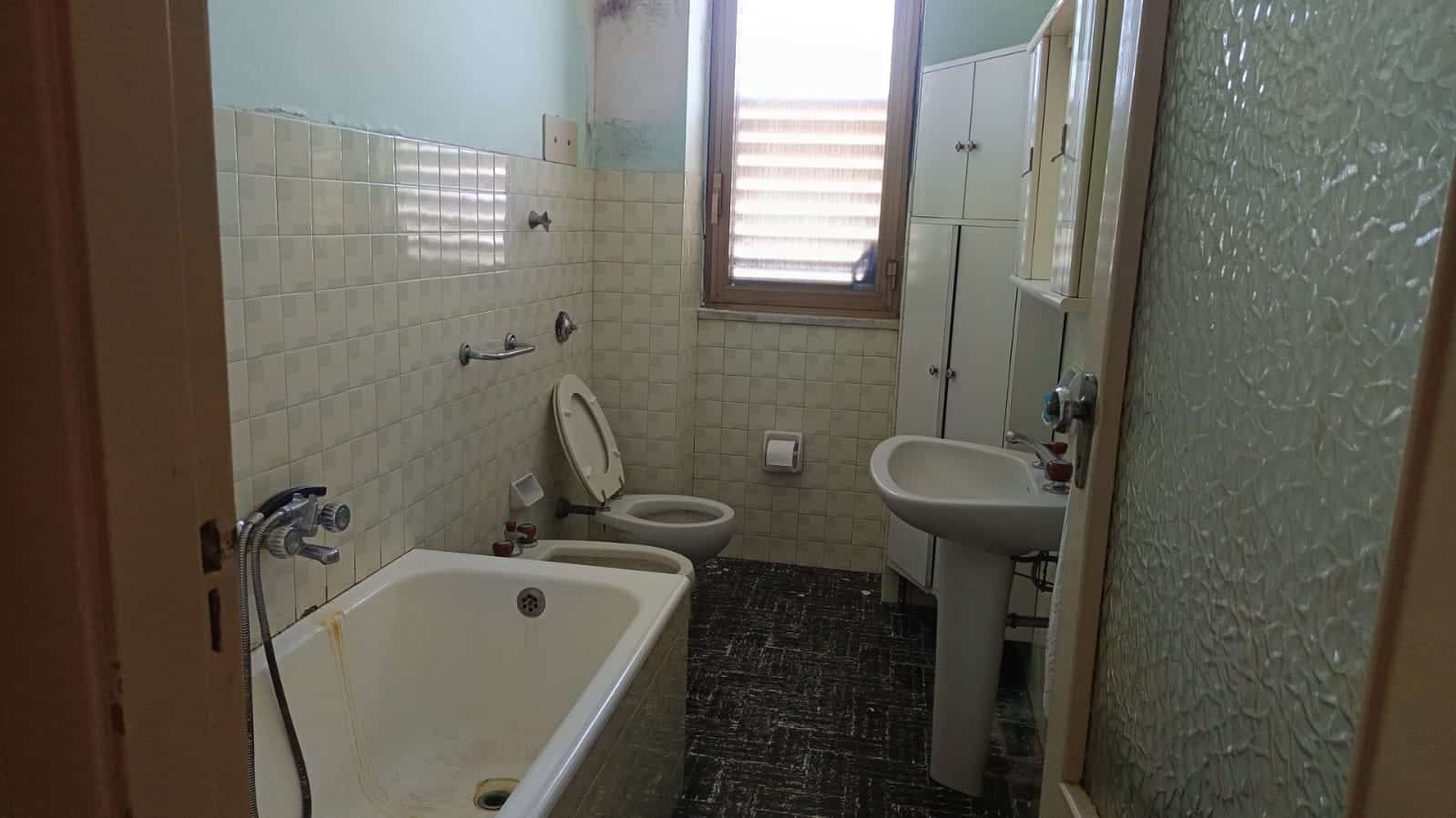 Bagno in appartamento con vasca, WC, bidet e lavabo, illuminato da finestra, in stile classico con piastrelle beige e pavimento a motivo scuro, zona lavanderia e stoccaggio integrati.