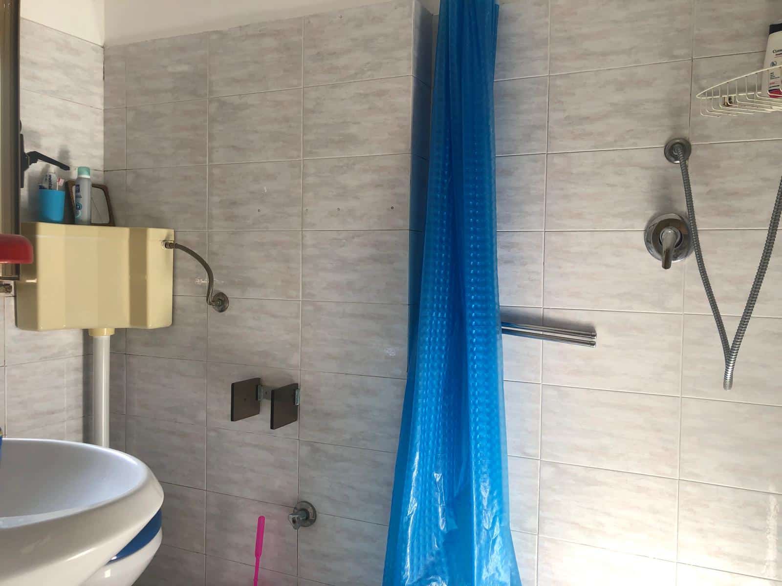 Lavabo e sedia da bagno in una cucina con rivestimento in piastrelle grigie e box doccia con tendina blu, accessori per la pulizia e rubinetti in vista, ambiente funzionale e pratico.