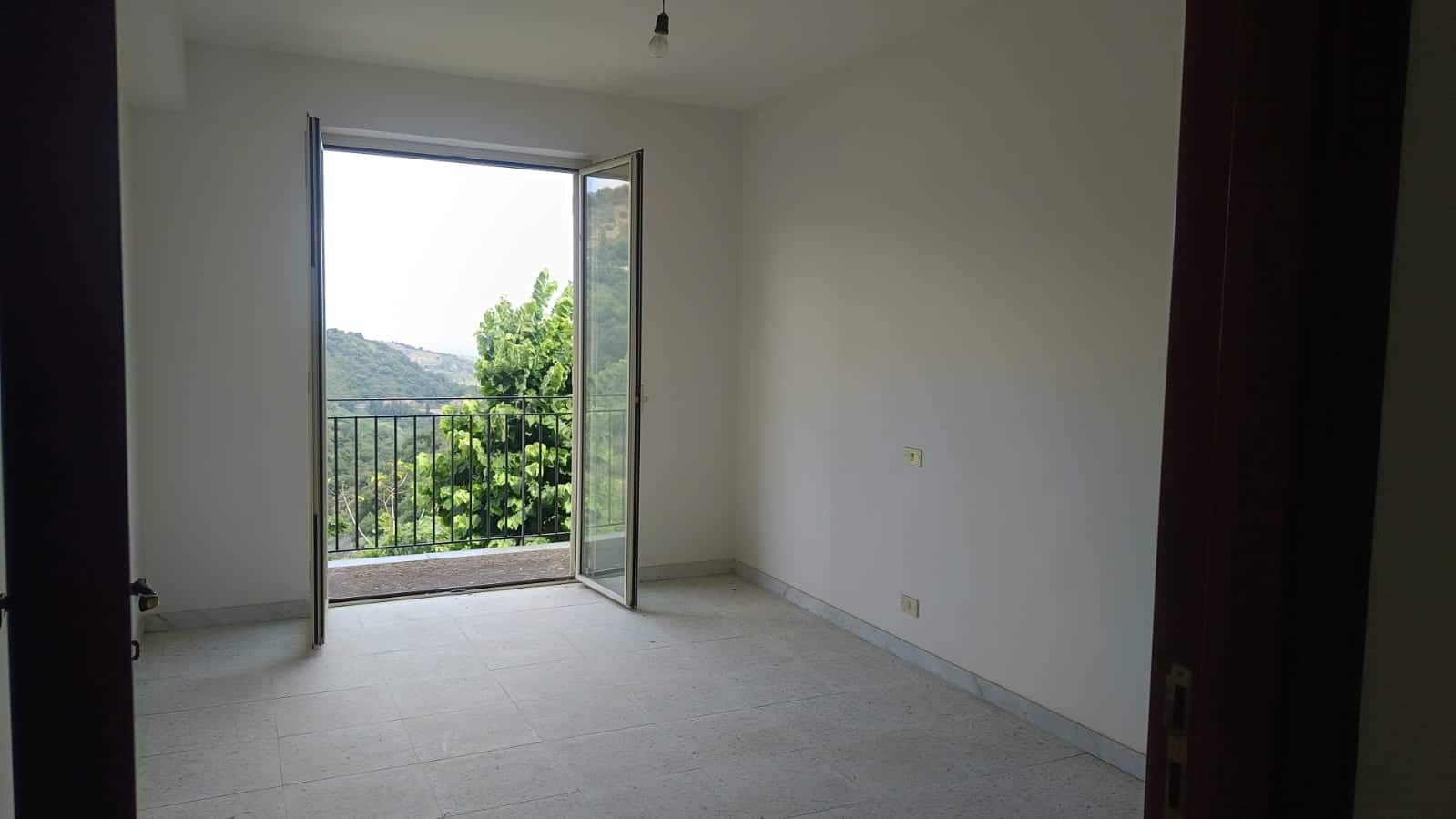 Vista di una stanza luminosa con porte-finestre che si affacciano su un balcone e una vista panoramica sulla natura e le montagne circostanti. Perfetta per investimenti immobiliari e case vacanza.