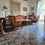 Antico salotto in stile classico con divani in pelle, mobili in legno e piastrelle decorate, ambiente luminoso con tende leggere, perfetto per soggiorni accoglienti e eleganti.