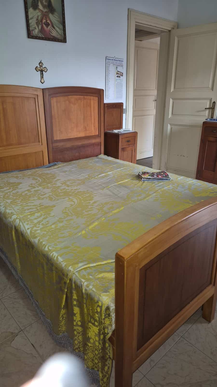 Letta, letto matrimoniale in legno con testiera classica in stile tradizionale, camera da letto accogliente e ben arredata, con arredamento in legno chiaro, pareti bianche e accessori religiosi, ideale per ambienti familiari e confortevoli.