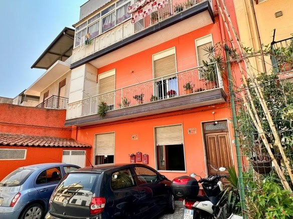 Vani in fiamma con azzurro e grigio, auto e moto parcheggiate davanti a un edificio residenziale color pesca con balconi e piante.