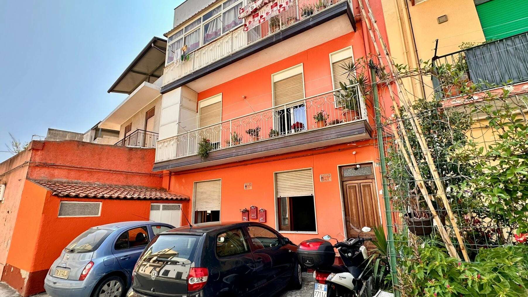 Vani in fiamma con azzurro e grigio, auto e moto parcheggiate davanti a un edificio residenziale color pesca con balconi e piante.