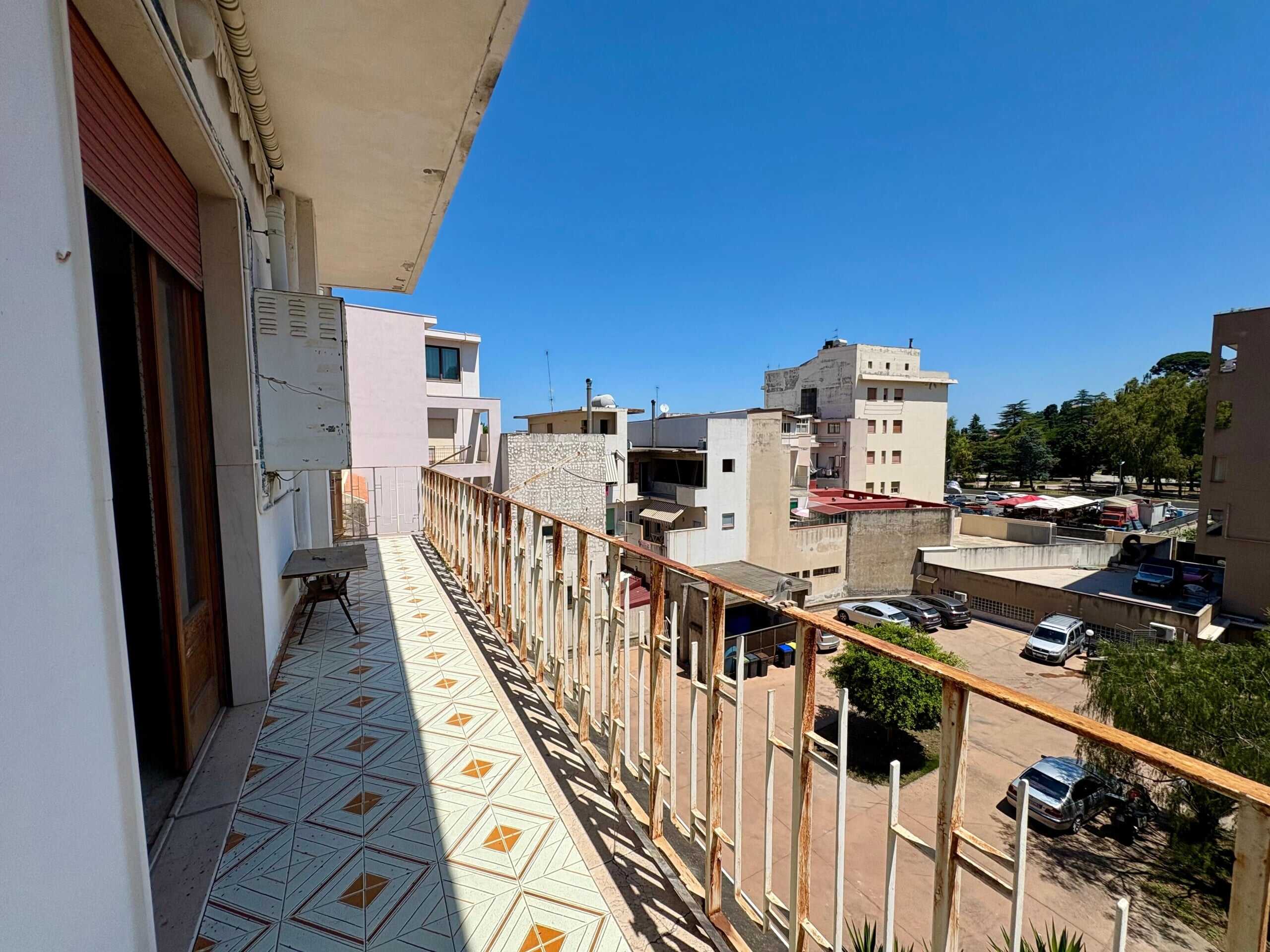 Balcone di un appartamento con pavimento in piastrelle decorative e vista sulla città, con edifici residenziali e un parcheggio. Ambiente luminoso e soleggiato.