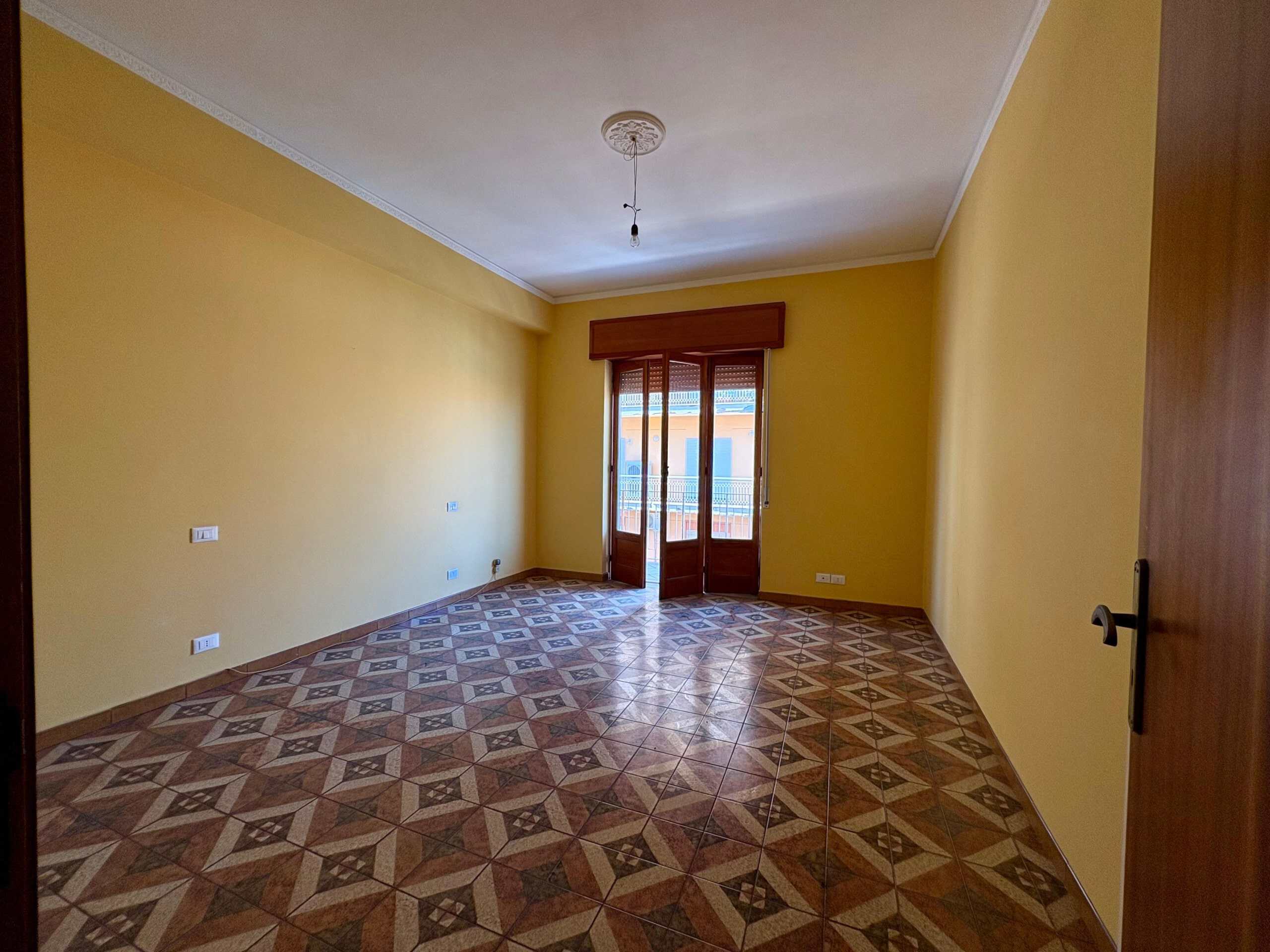 Spazio abitativo luminoso con pareti gialle, pavimento in piastrelle geometriche e balcone con accesso tramite portafinestra in legno.