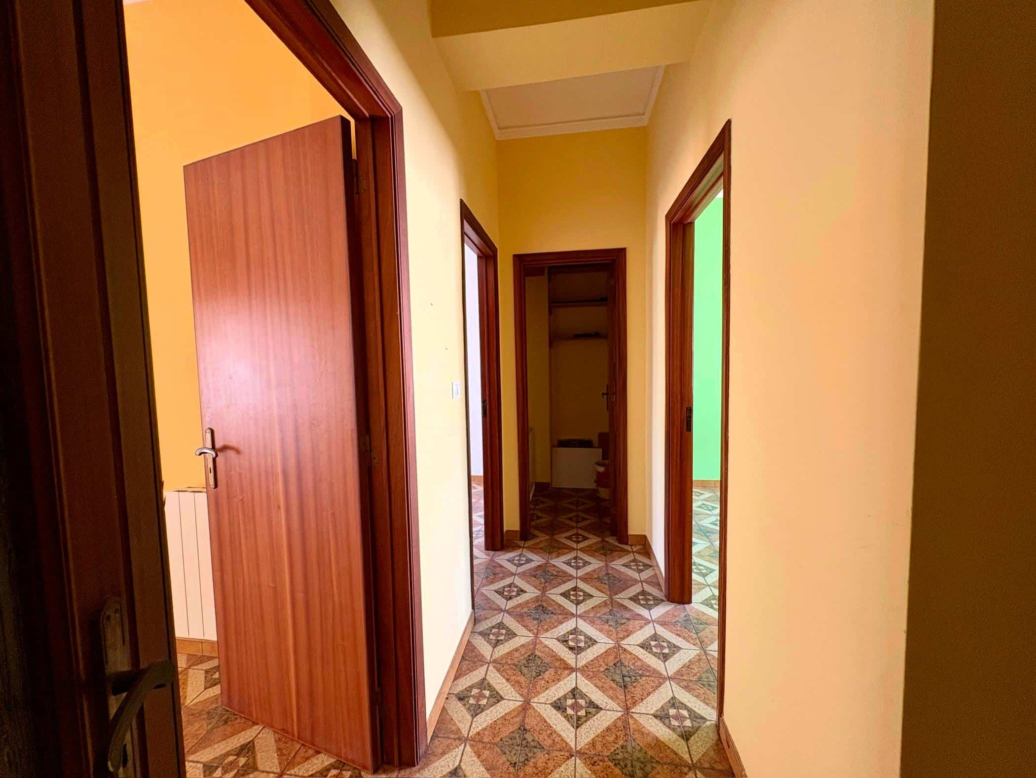 Porta d'ingresso in legno chiaro, corridoio con pareti gialle e pavimento in piastrelle colorate, con accesso a diverse stanze, in un appartamento in vendita o in affitto.