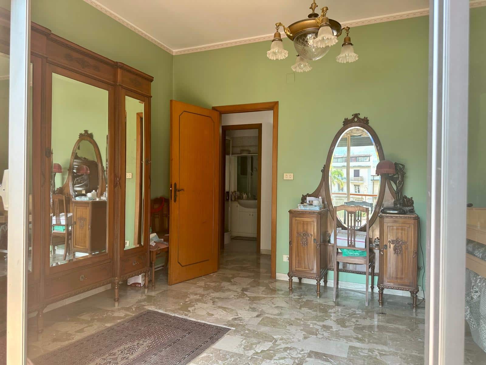 Antico mobile in stile provenzale e grande armadio in legno, ingresso con pareti verdi, pavimento in marmo beige, dettagli in stile classico, lampadario vintage, specchio ovale con mobiletto in legno, vista verso l’interno dell’appartamento, arredamento tradizionale, ambiente elegante e accogliente, atmosfera calda e nostalgica.