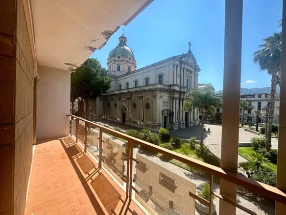 Vista panoramica sulla chiesa storica di Santa Maria delle Grazie a Napoli, visibile dal balcone di un appartamento, con la cupola e il cielo sereno sullo sfondo, tra palme e edifici eleganti.