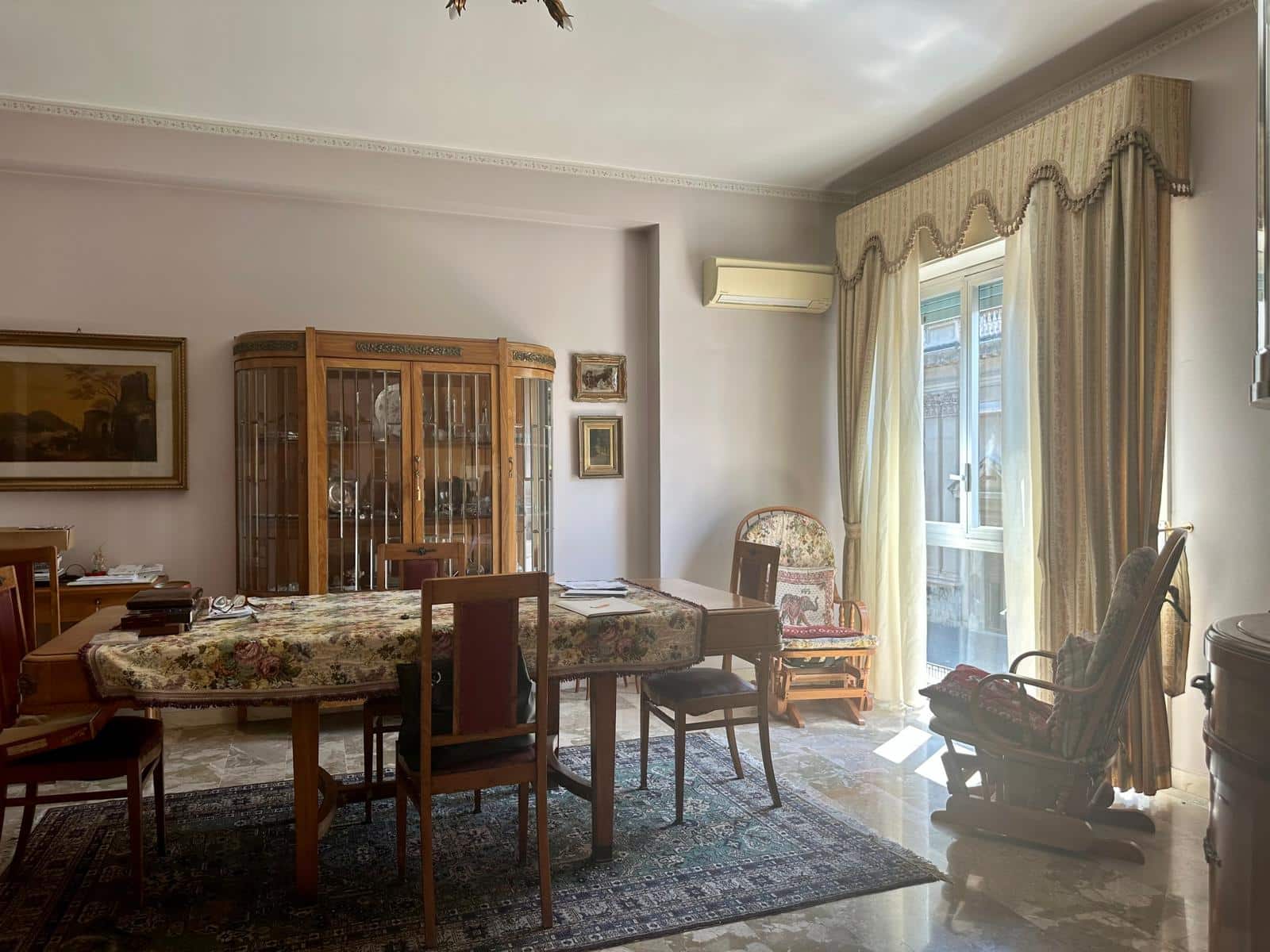 Vasta stanza da pranzo con mobili d'epoca, tappeto orientale e grande finestra con tende ornate, ambiente elegante e accogliente.