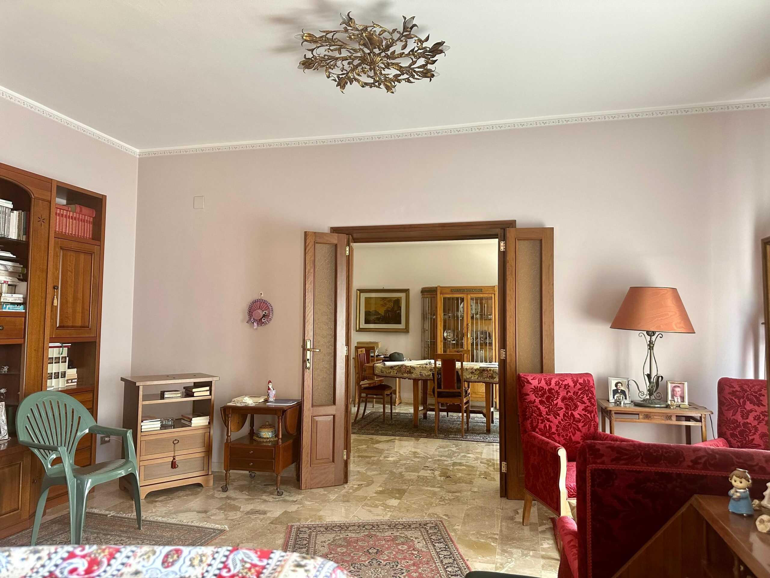 Soggiorno elegante con pareti chiare, mobili in legno, sedie e divani imbottiti rossi, studiolo con lampada e cornici, ambiente accogliente e ben arredato.