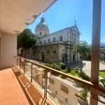 Vista dal balcone con affaccio sulla chiesa storica e la piazza centrale di Catania, con cielo sereno e palme ornamentali in un contesto urbano elegante.