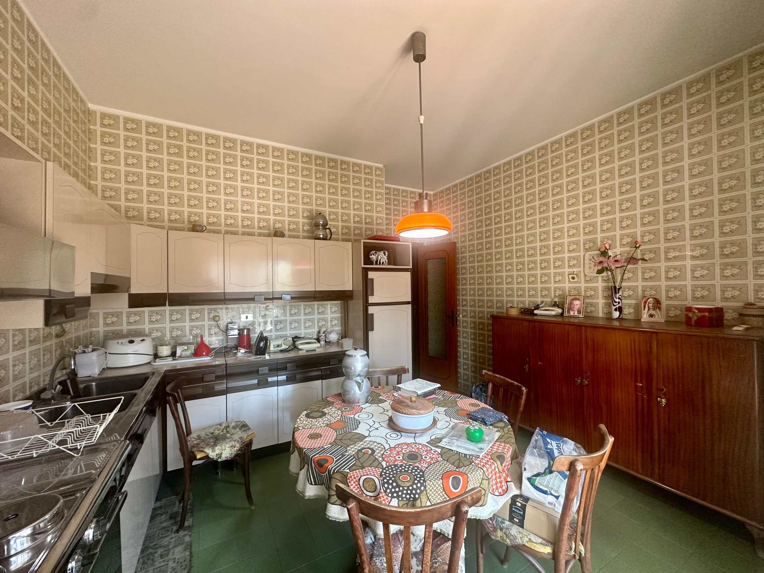 Vecchia cucina in stile vintage con mobili in legno e piastrelle retrò, tavolo da pranzo con tovaglia colorata, oggetti di uso quotidiano e decorazioni domestiche, illuminazione con lampada arancione, ambiente accogliente e caratteristico.