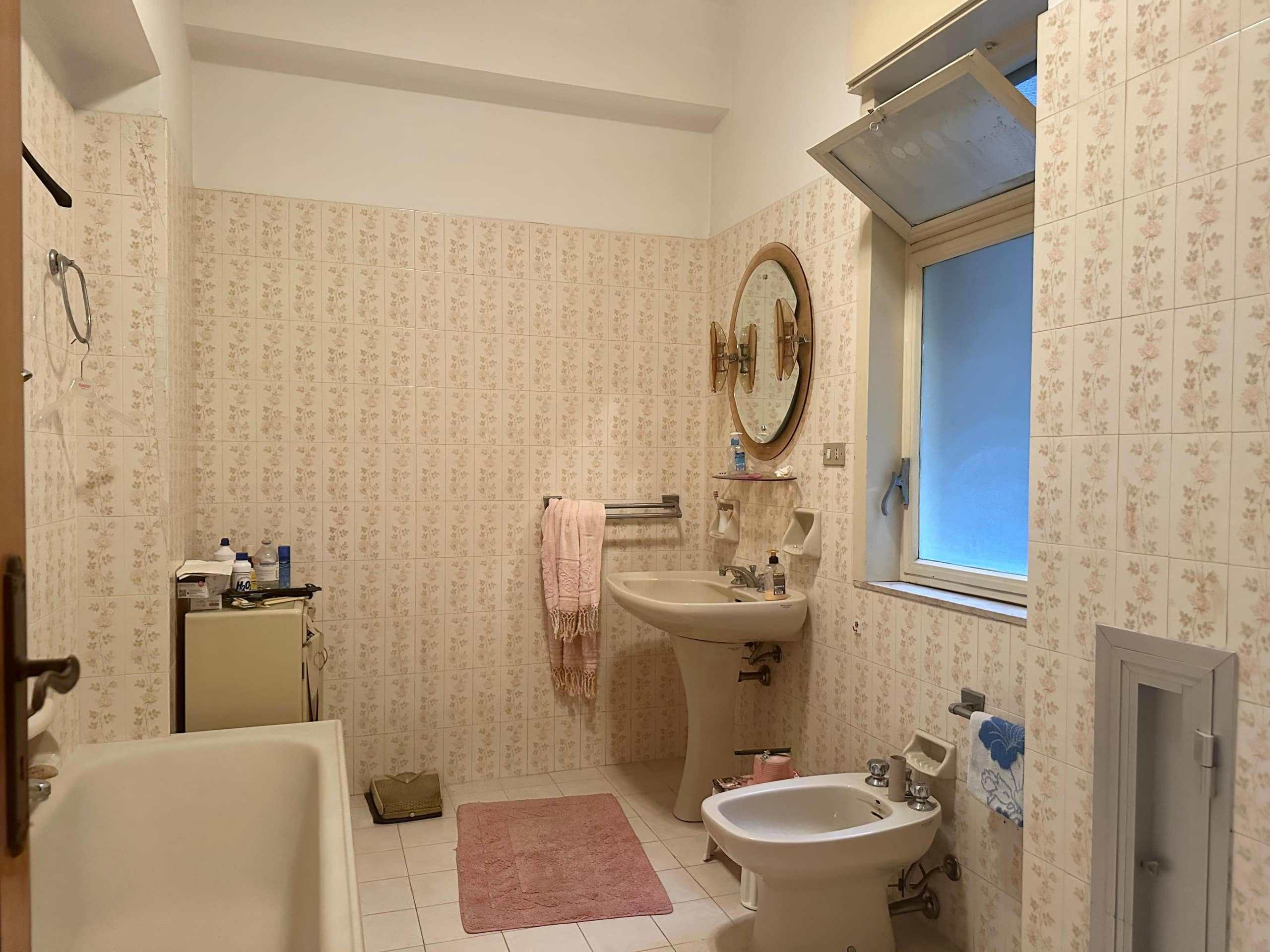 Ampio bagno con mosaico floreale, lavabo con mobile in stile vintage, bidet, vasca, finestra panoramica, accessori bagno e armadietto di servizio, ideale per famiglie o case d'epoca.