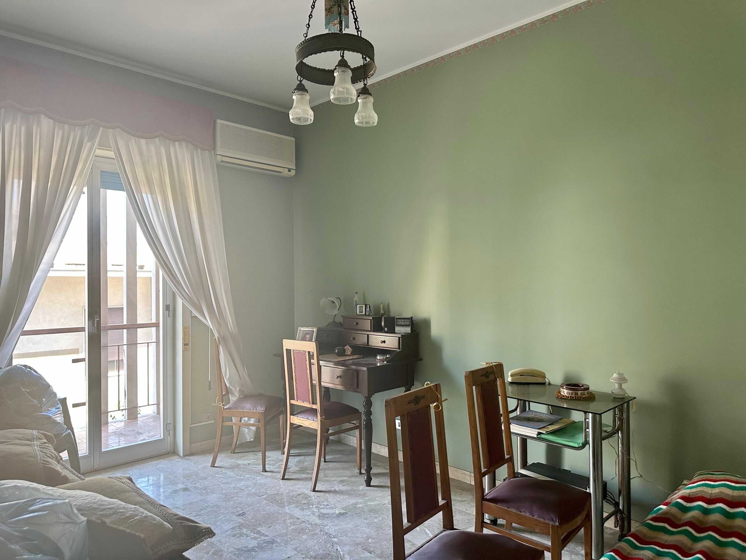 Foto di un soggiorno con parete verde, grande balcone con tende bianche, mobili classici, tavolo con sedie, aria condizionata e lampadario antico, ambiente accogliente e luminoso.