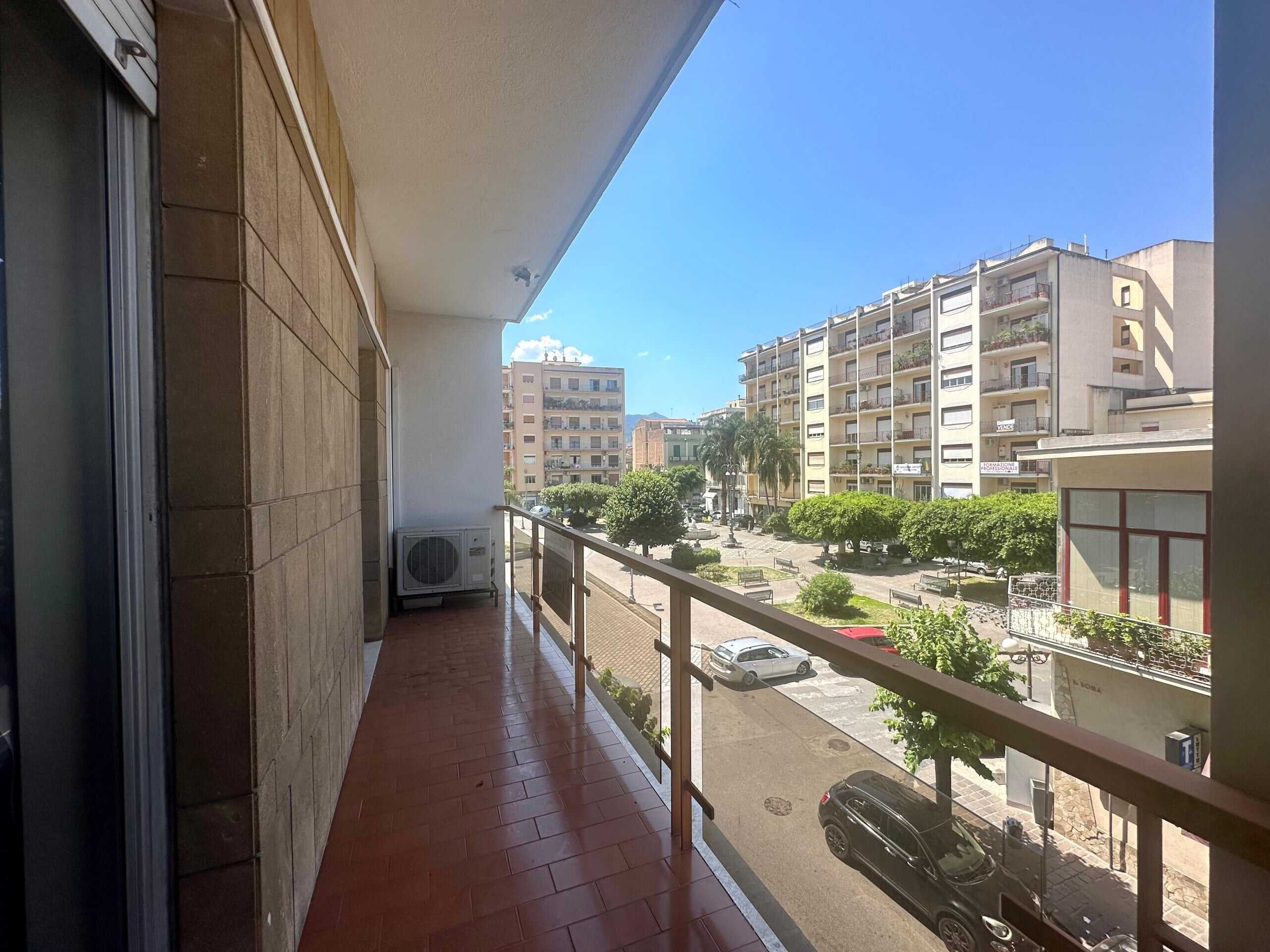 Balcone di appartamento in zona urbana, vista su parco e palazzi residenziali, pavimentato in terracotta, con aria condizionata esterna, ideale per vivere in città.