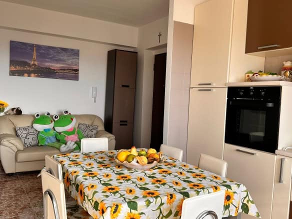 Finestra di un appartamento abitabile con cucina e zona pranzo, decorata con tende a girasole, oggetti d'arredo colorati e bambole giganti di rana, ambiente accogliente e funzionale, ideale per vivere a NextCasa.