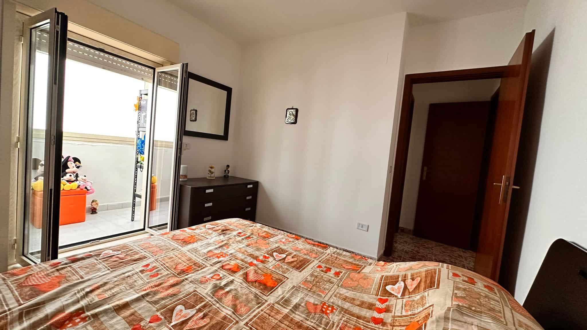 Stanza da letto luminosa con balcone, letto matrimoniale con biancheria a tema cuore, armadio, pareti bianche e decorazioni semplici, ideale per vivere in comfort e relax.