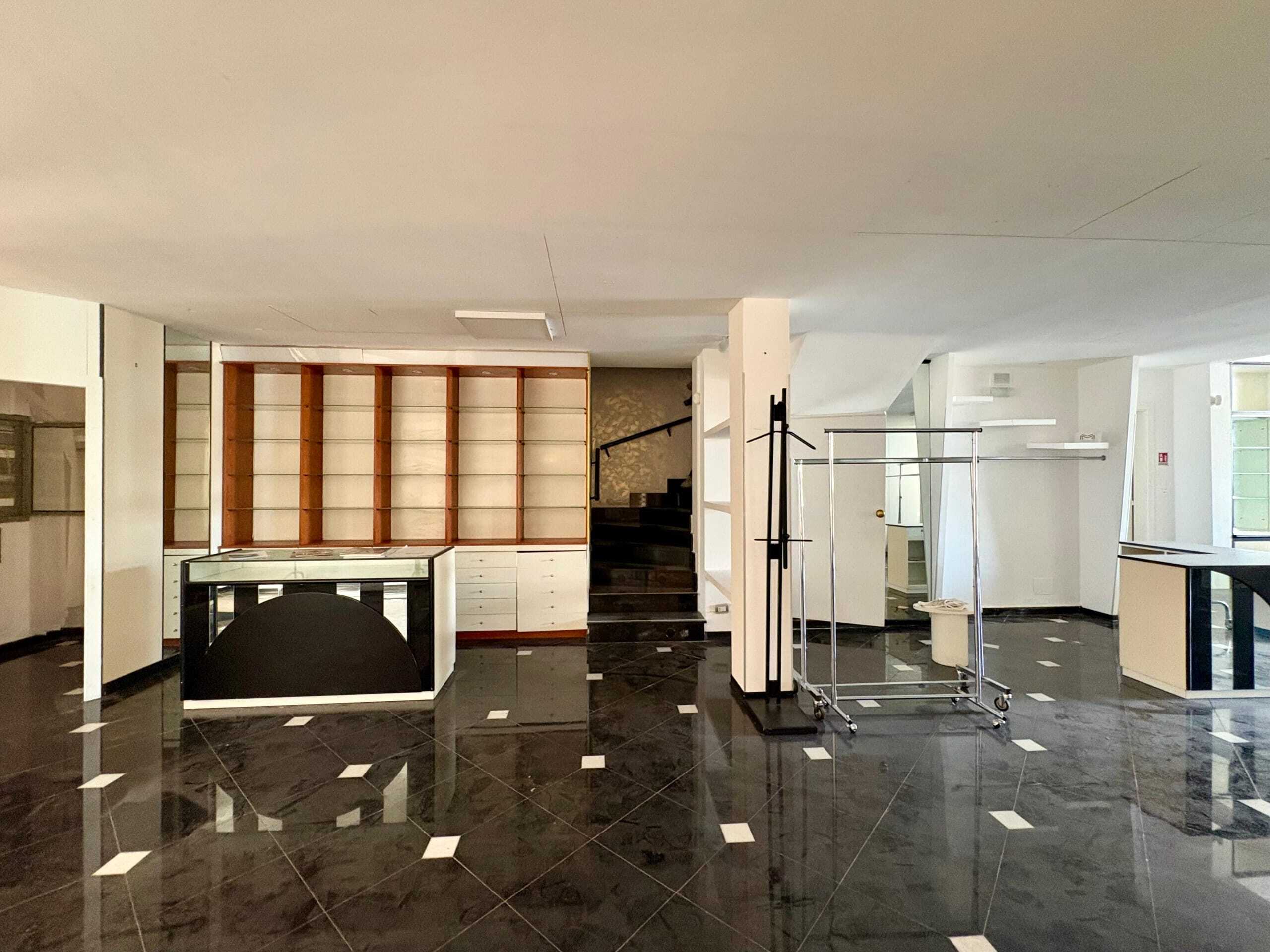 Juxtaposto di mobili da ufficio e scaffalature vuote in uno spazio commerciale o showroom. Pavimento in piastrelle nere lucide con inserti bianchi, pareti bianche e finestre che permettono l'ingresso di luce naturale.