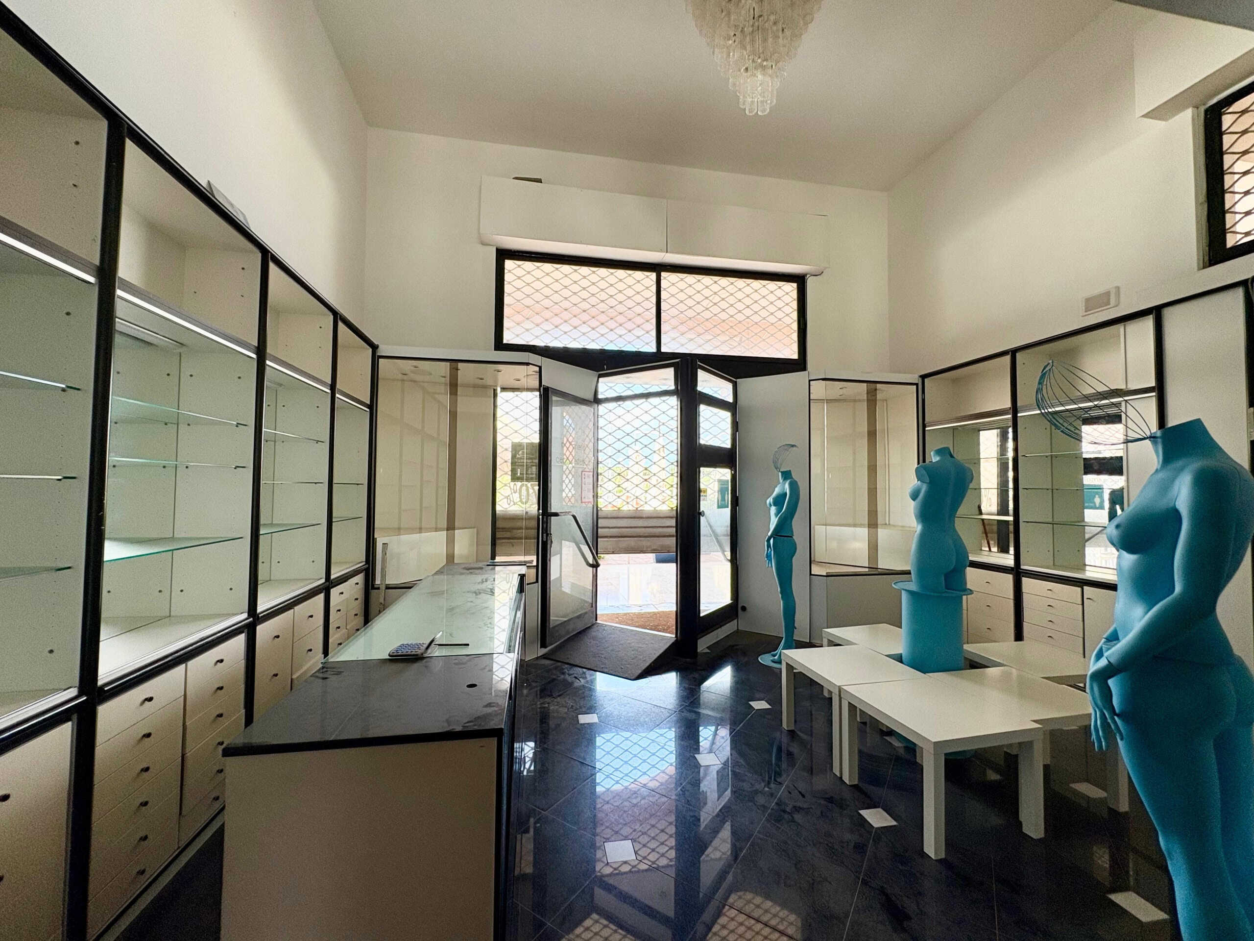 Vetrina commerciale vuota con manichini blu in esposizione, interior design moderno, negozio di abbigliamento o showroom, pavimento in piastrelle nere lucide, grandi finestre con grate di sicurezza, spazio luminoso e elegante.