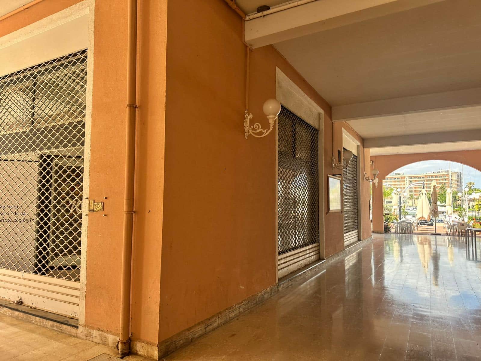 Prospettiva di un portico con pavimento in marmo e pareti color terracotta, con finestre e porte con grate metalliche e luci vintage, che conduce a un'area esterna con tavoli e sedie sotto ombrelloni, in un complesso residenziale.
