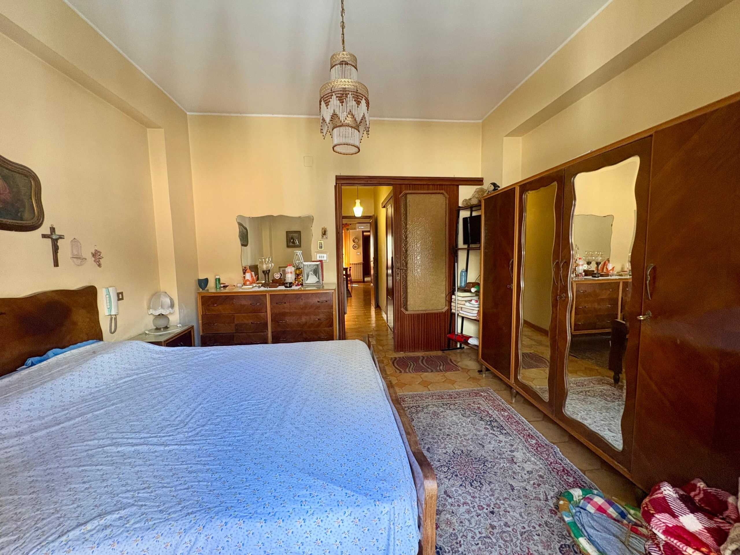 Letto matrimoniale in camera da letto con mobili vintage e armadio con specchio. Ambientazione accogliente con dettagli classici e decorazioni religiose, ideale per appartamenti di charme e residenze storiche.