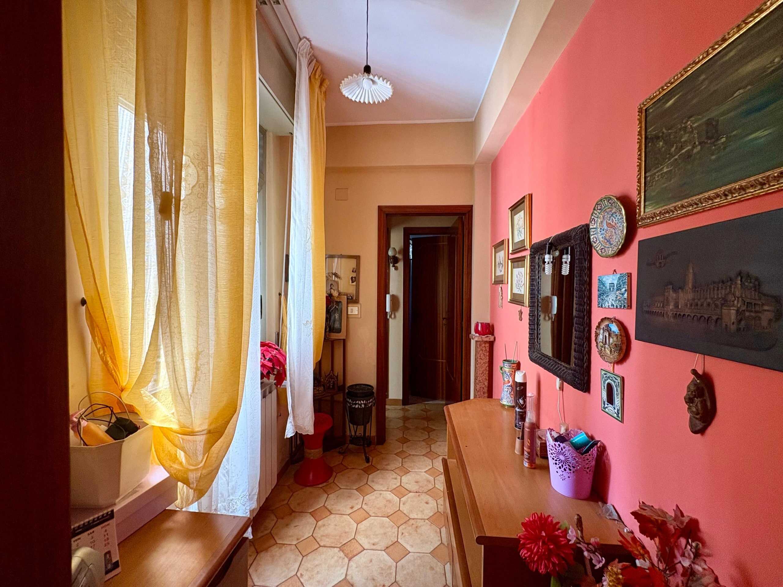 Locale d'ingresso con pareti colorate, decorato con quadri e specchio, finestra con tende gialle, arredi in legno e oggetti decorativi, atmosfera accogliente e calda.