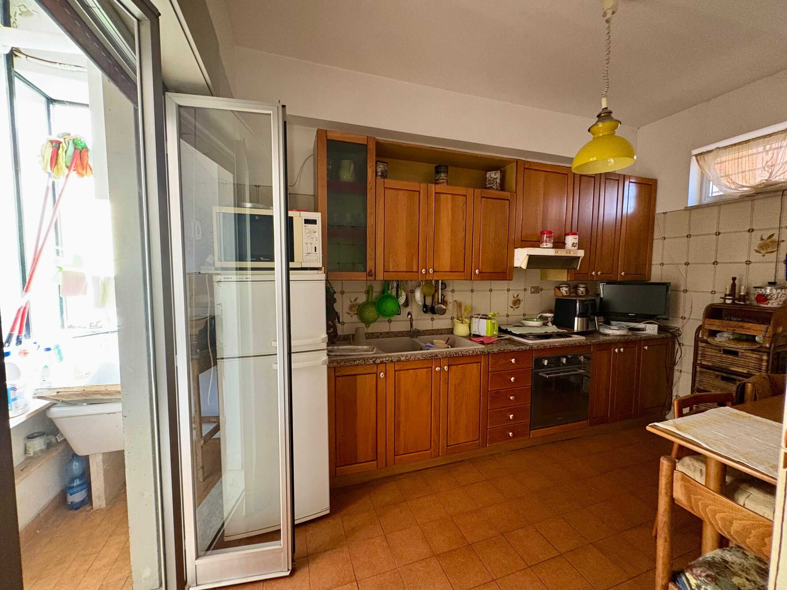 Ripostiglio e cucina abitabile con mobili in legno, forno, frigorifero, stoviglie e accessori, ambiente funzionale e accogliente, zona giorno luminosa con finestra, arredamento pratico e dettagli casalinghi.