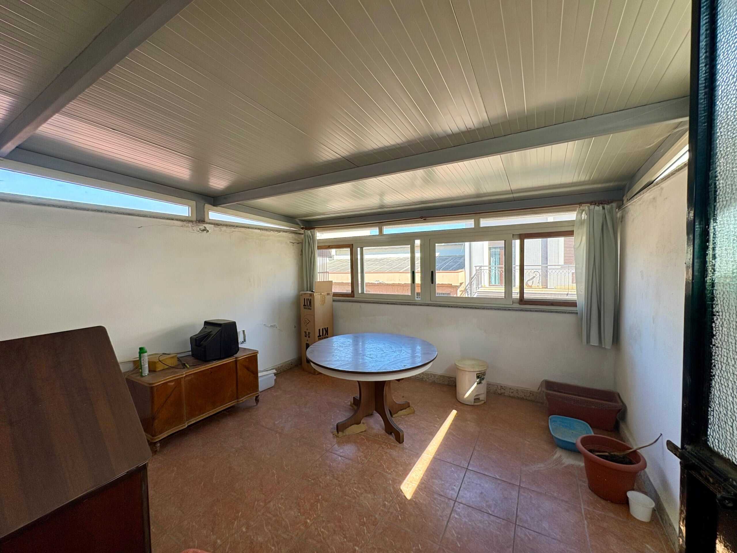 Scala: interni di un terrazzo o balcone abitabile con pareti bianche, grande finestra a tutta lunghezza, arredato con tavolo rotondo, mobile in legno, e piante in vaso, luce naturale.
