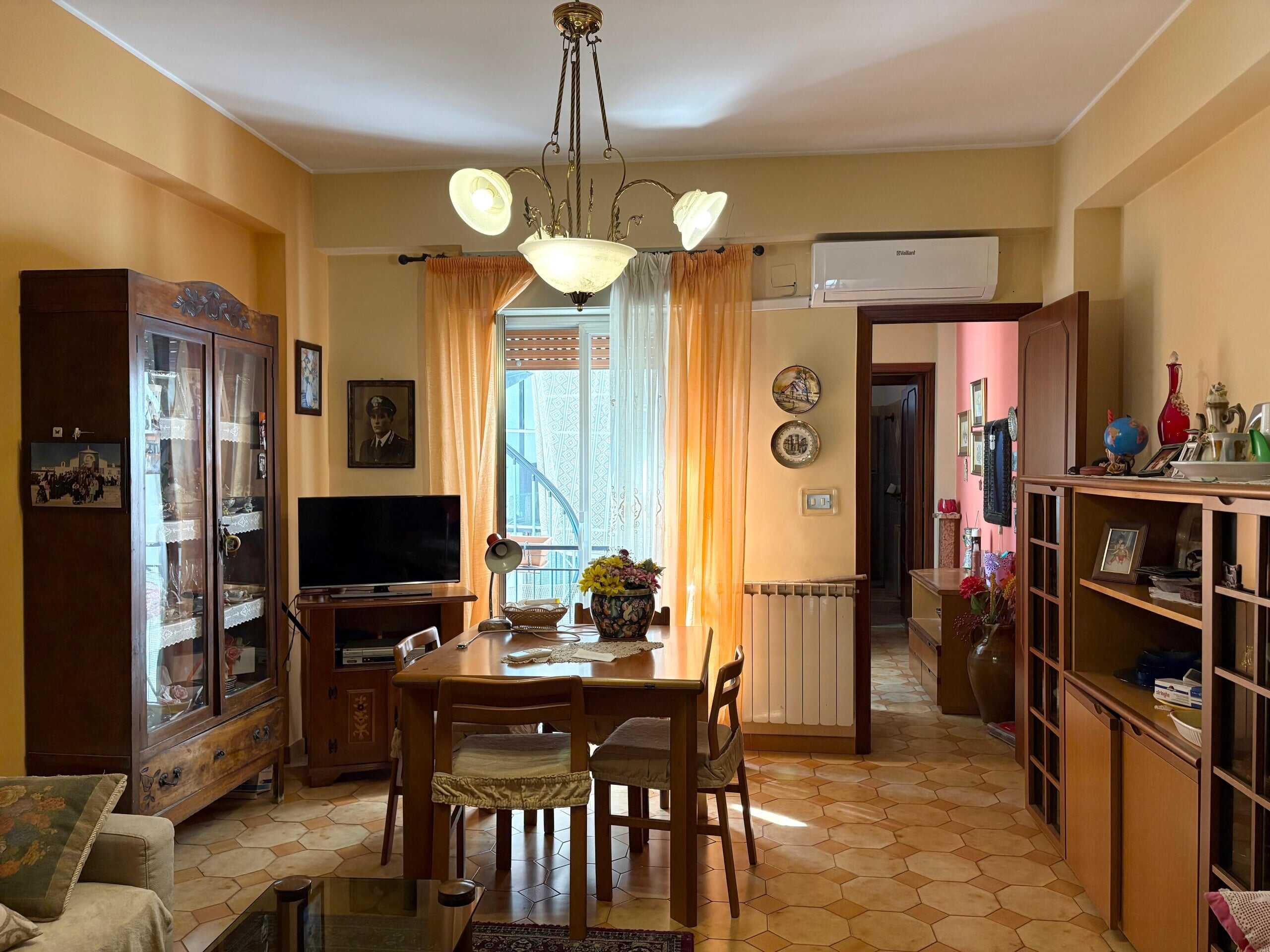 Soggiorno spazioso con arredi in legno, tavolo da pranzo, televisore e decorazioni vintage, illuminato da lampadario centrale, con accesso a balcone e aria condizionata, ideale per vivere con comfort.