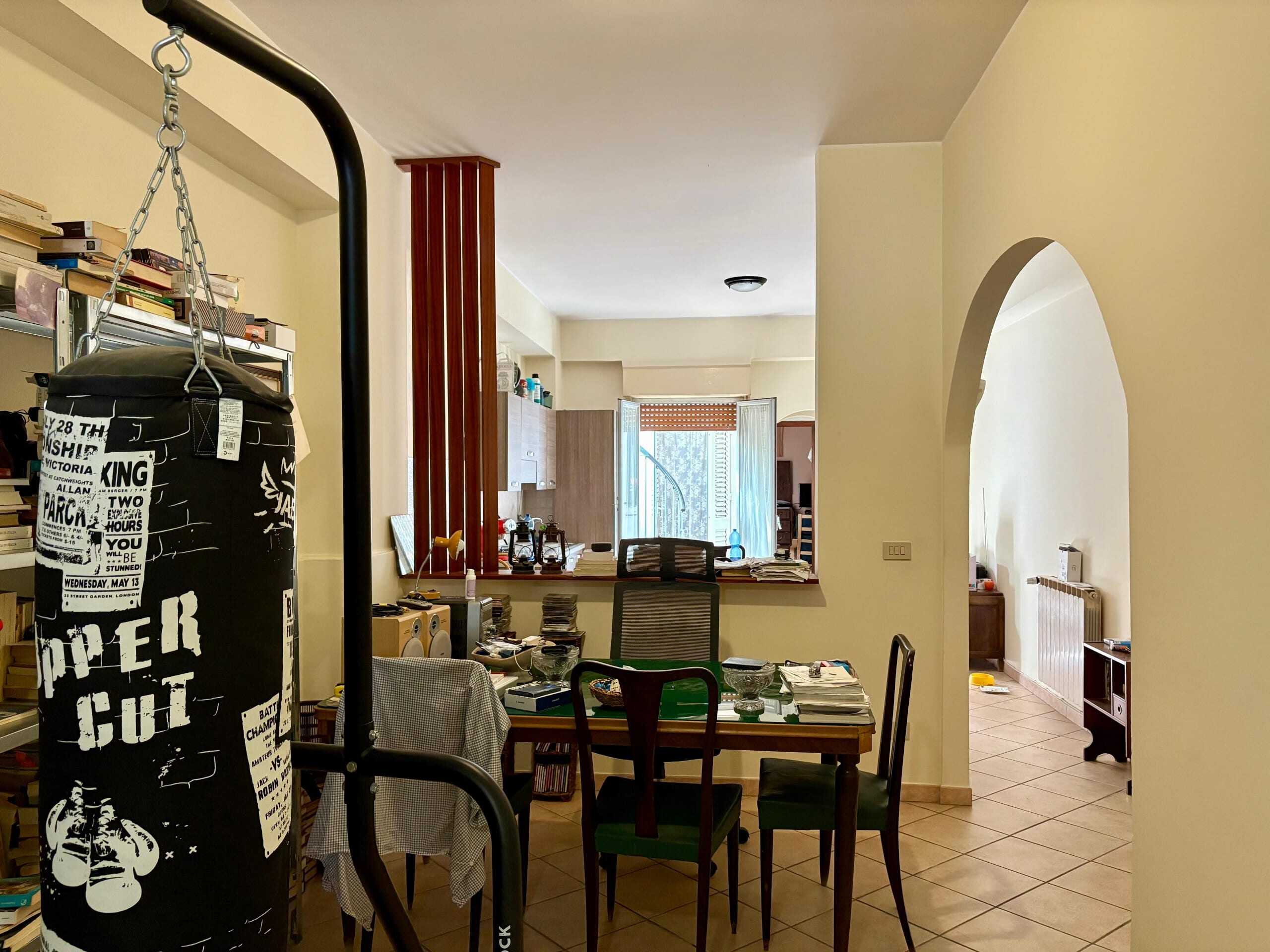 Punching bag e zona living con tavolo e sedie, ambiente domestico accogliente e disordinato in appartamento, spazio interno con arredi e libri, luminoso con finestra aperta.