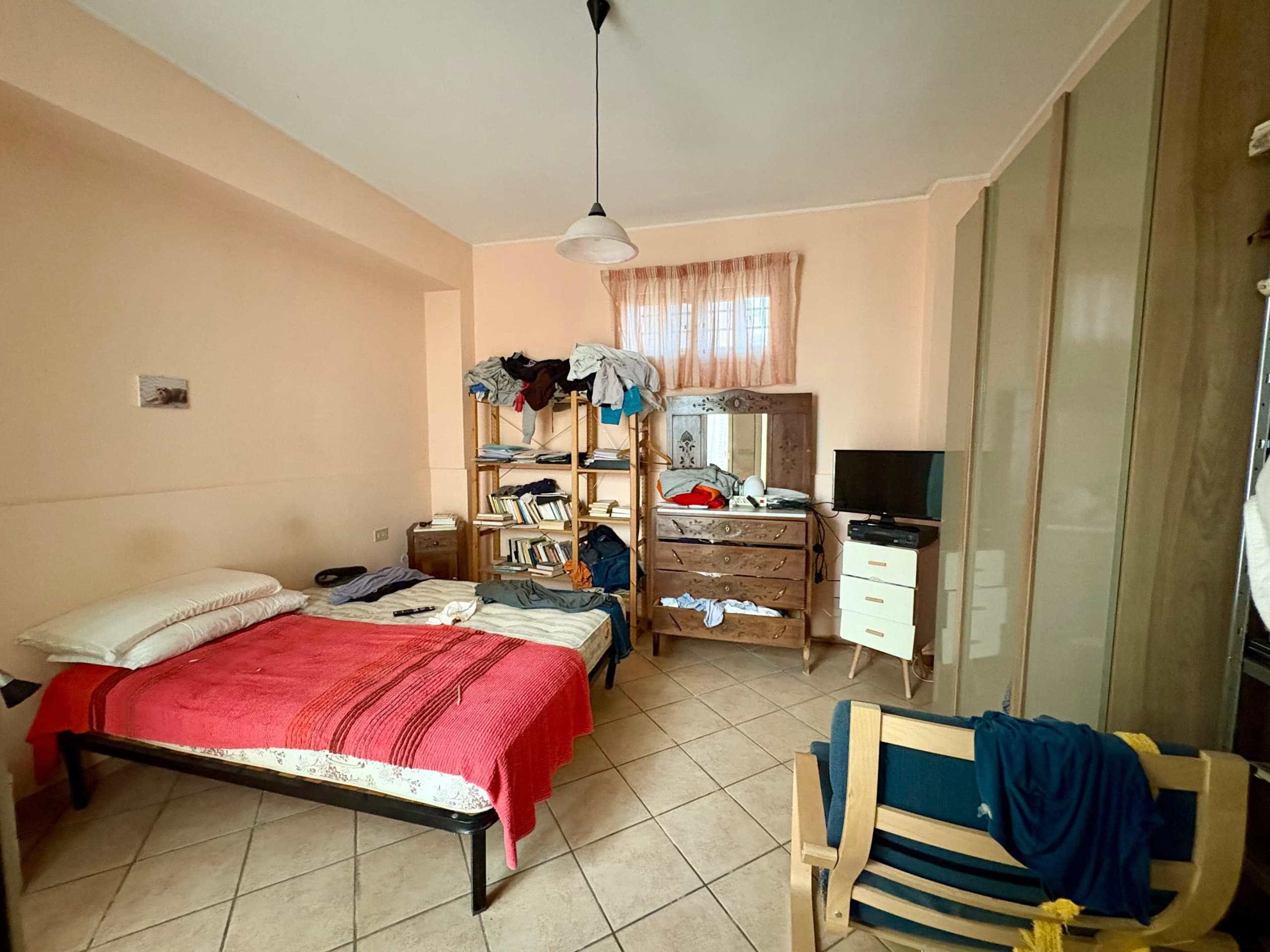 Letto matrimoniale disordinato in stanza con armadio e mobile tv, atmosfera informale e vissuta, dettagli di arredo rustico e disordine, spazio abitativo quotidiano, soluzione abitativa con stile semplice e pratico.