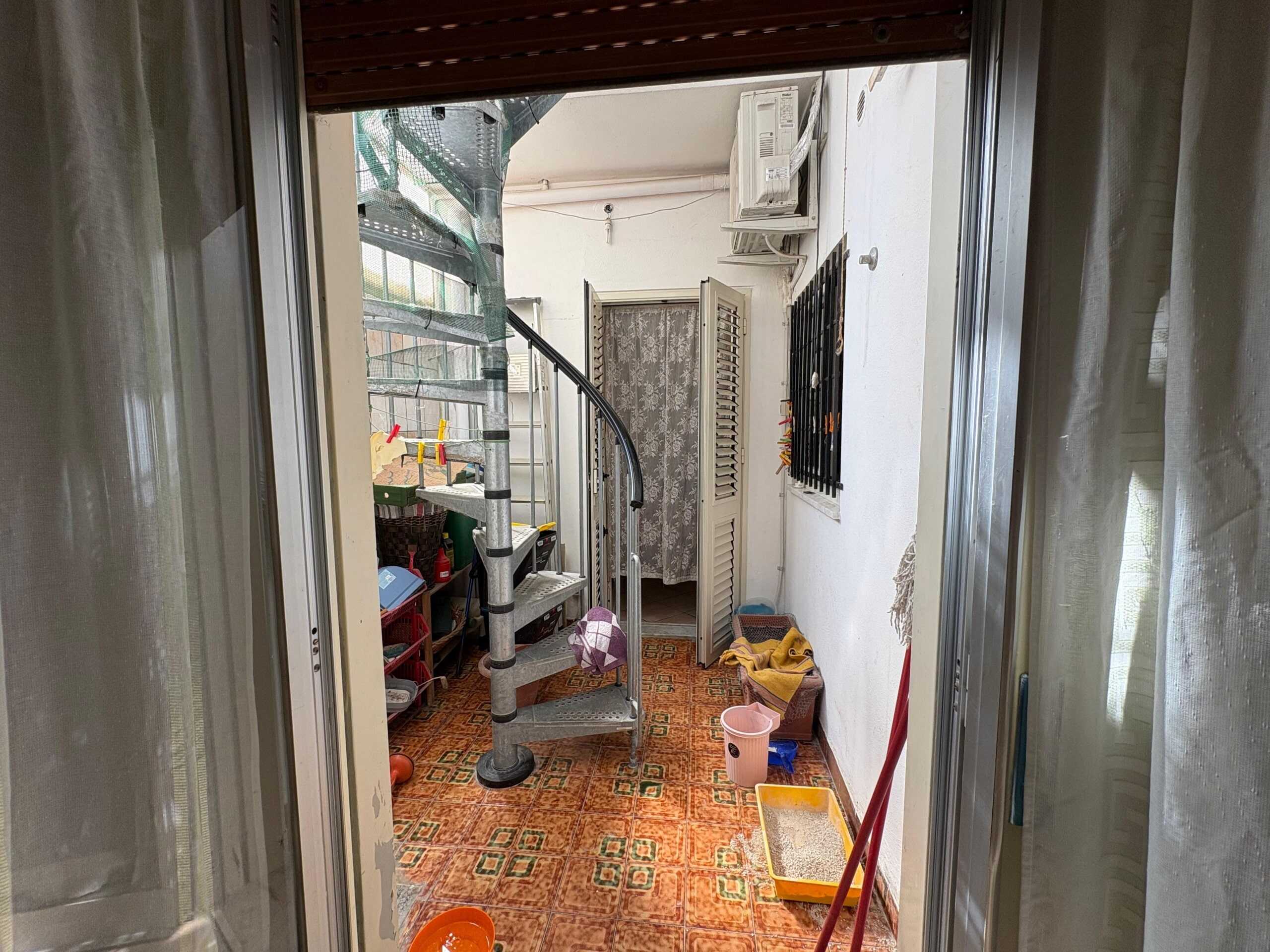 Spazio esterno di un appartamento con scala a chiocciola, arredi e oggetti per la casa, pavimento in piastrelle 1970, elementi di sicurezza come grate alle finestre.