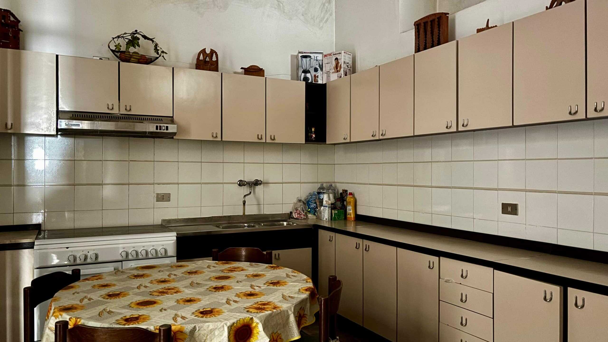Lavagna cucina con ante beige, piano di lavoro nero e pareti a piastrelle bianche, tavolo rotondo con tovaglia a girasole, ambiente abitabile e funzionale.