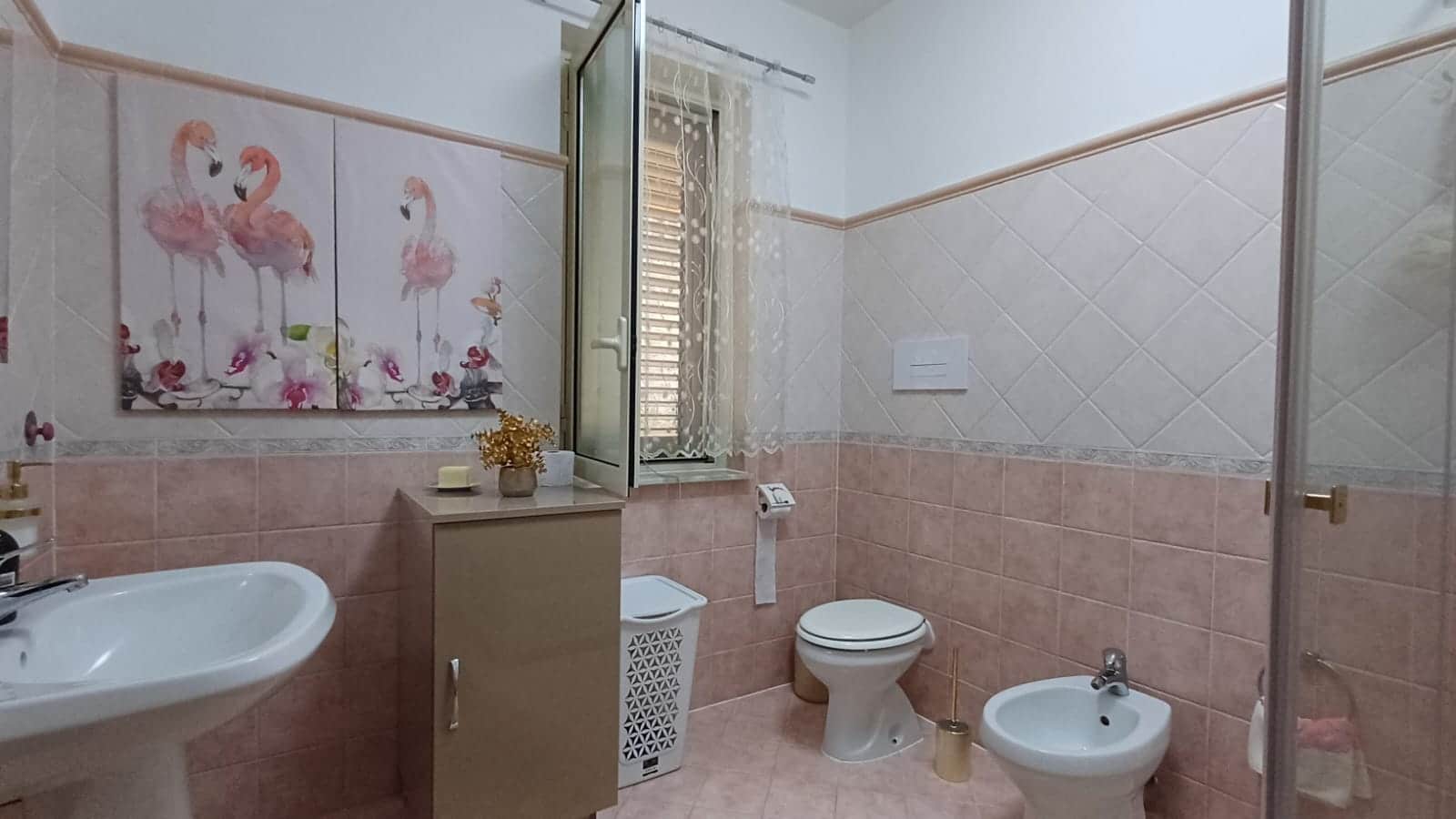 Bagno con piastrelle rosa e cream, lavabo, WC, bidet, finestra con tende decorative, decorazioni flamingo, stile classico e accogliente, ideale per appartamenti e case vacanza.