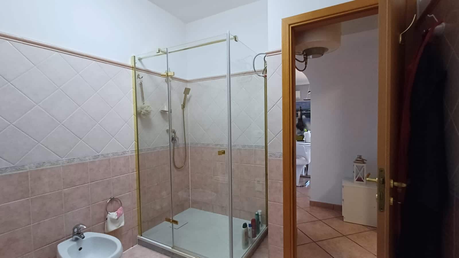 Box doccia in vetro con apertura a battente e rivestimento in piastrelle beige e bianco, ambiente bagno funzionale e semplice, con porta in legno e decorazioni eleganti, ideale per appartamenti NextCasa.