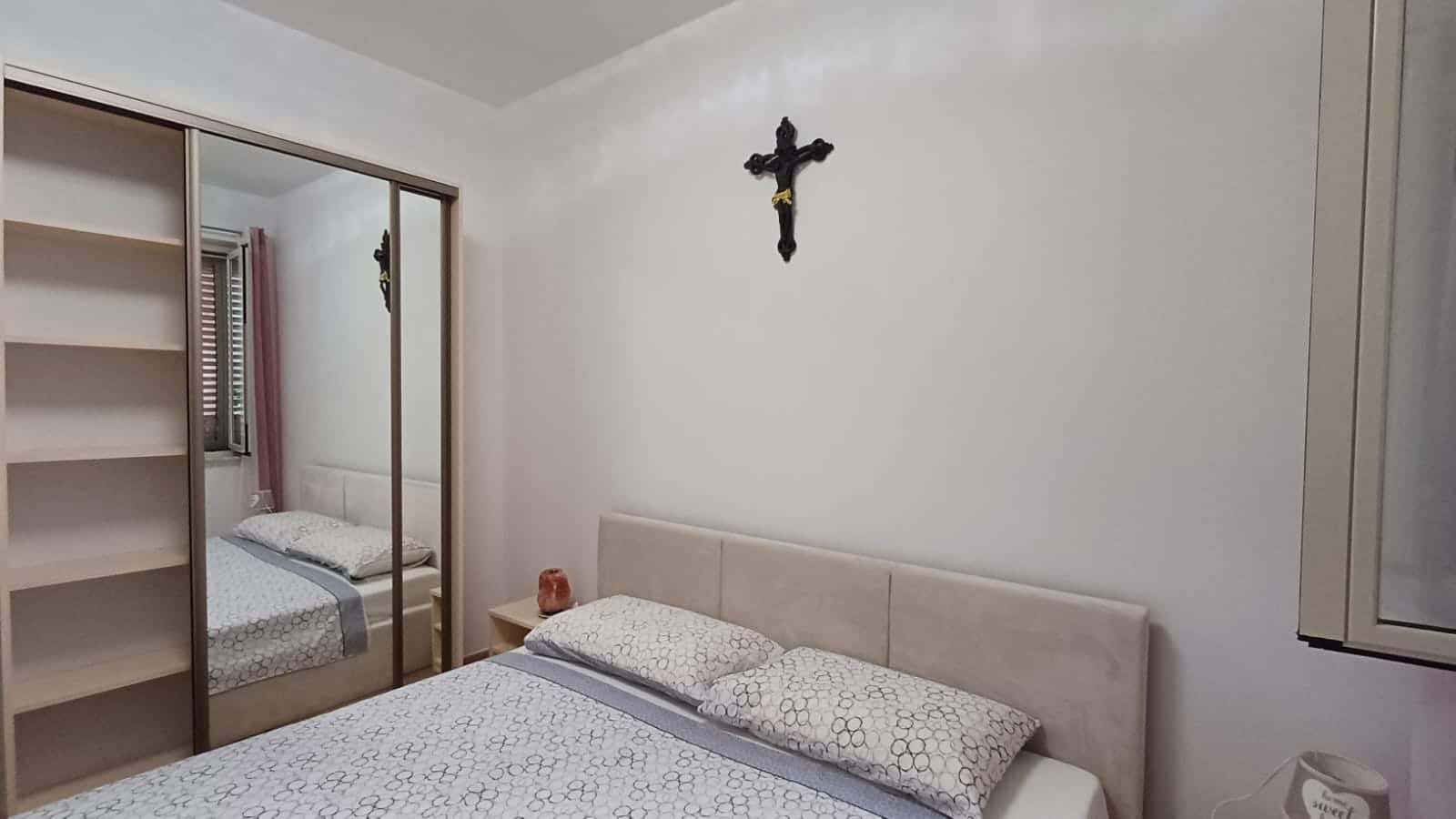 Camera da letto con letto matrimoniale, armadio a specchio e decorazione religiosa sulla parete, arredamenti moderni, ambiente luminoso, ideale per acquisto o affitto immobiliare.