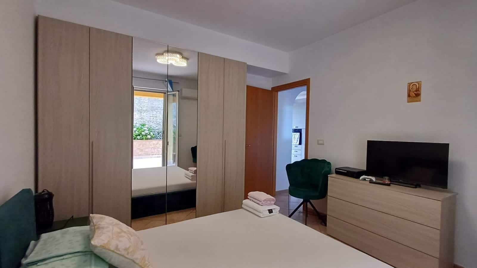 Armadio a muro con ante in legno e specchio, soggiorno elegante con TV e arredi moderni, camera da letto luminosa, porta che conduce al balcone con vista sulla natura, soluzione abitativa confortevole.
