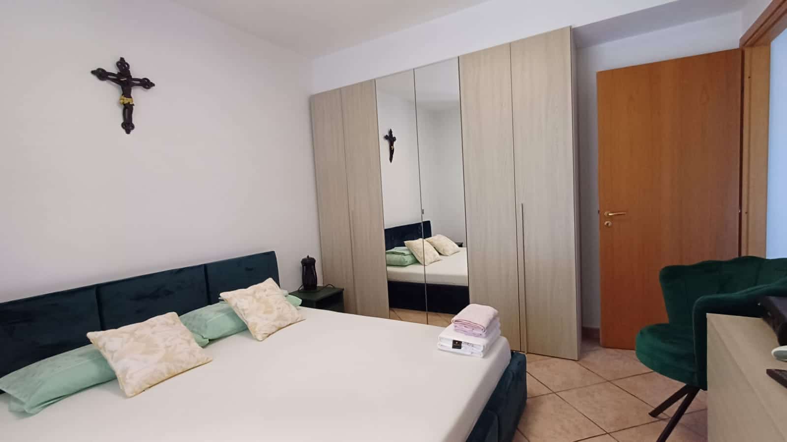 Letto matrimoniale con cuscini beige e verdi, arredi in stile semplice, armadio a ante scorrevoli con specchio, pareti bianche, decorazioni religiose, ambiente accogliente e funzionale.