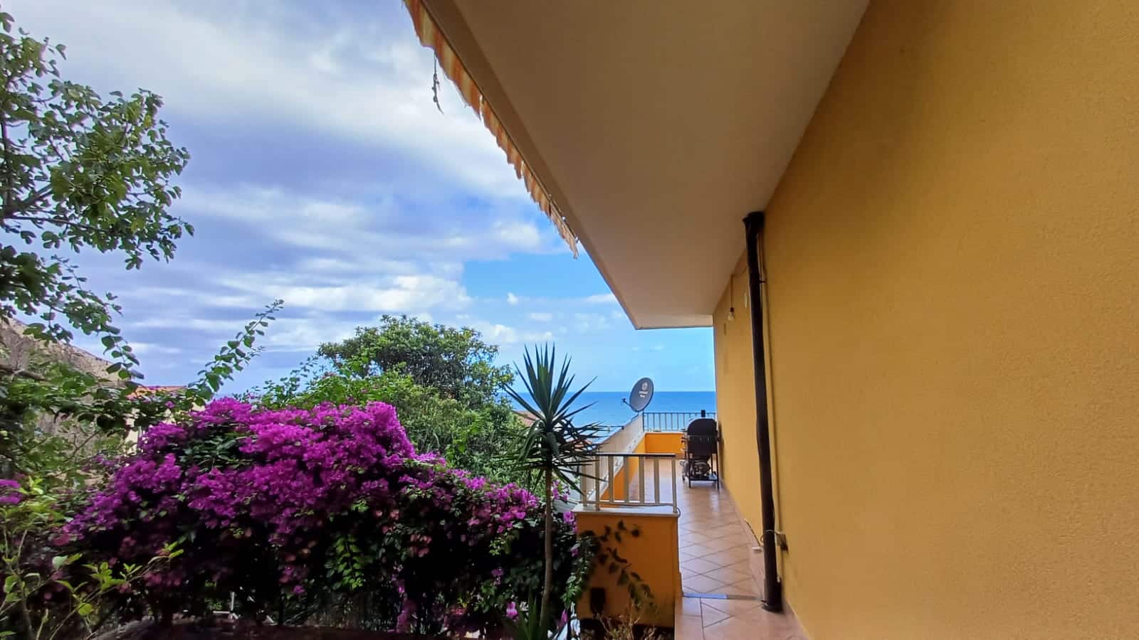 Balcone con vista mare, fiori colorati e cielo nuvoloso, appartamento in affitto o vendita vicino al mare, spazio esterno con piante tropicali, soggiorno con vista panoramica, NextCasa immobiliare, proprietà in Italia, case sul mare, ville con balcone, immobili in affitto con vista mare.