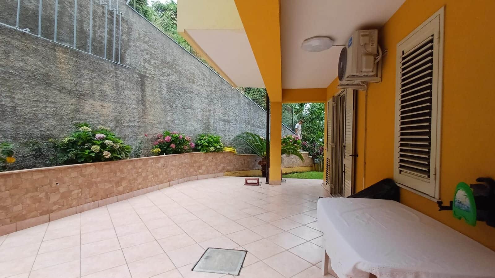 Balcone abitabile con fioriere, tettoia e aria condizionata, vista su giardino e mura di cinta, ideale come spazio esterno per relax e convivialità.