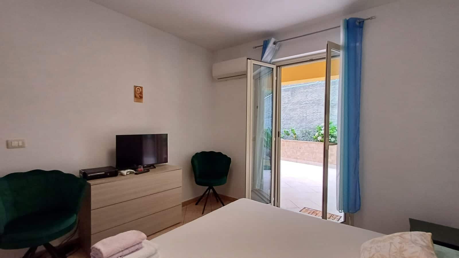 Camera da letto con porta finestra che dà su terrazza, aria condizionata, mobili moderni, TV, decorazioni semplici, tende blu e frigo con vista sul giardino, ambiente confortevole e luminoso.