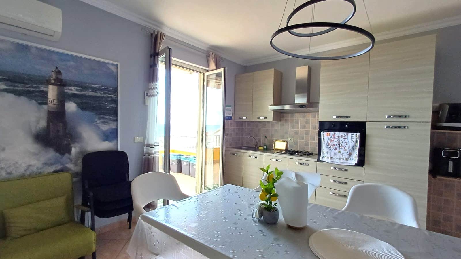 Locale cucina soggiorno luminoso con finestra e balcone, arredato con cucine moderne in tonalità beige, tavolo con tovaglia, sedie e decorazioni floreali, ideale per vacanze al mare.
