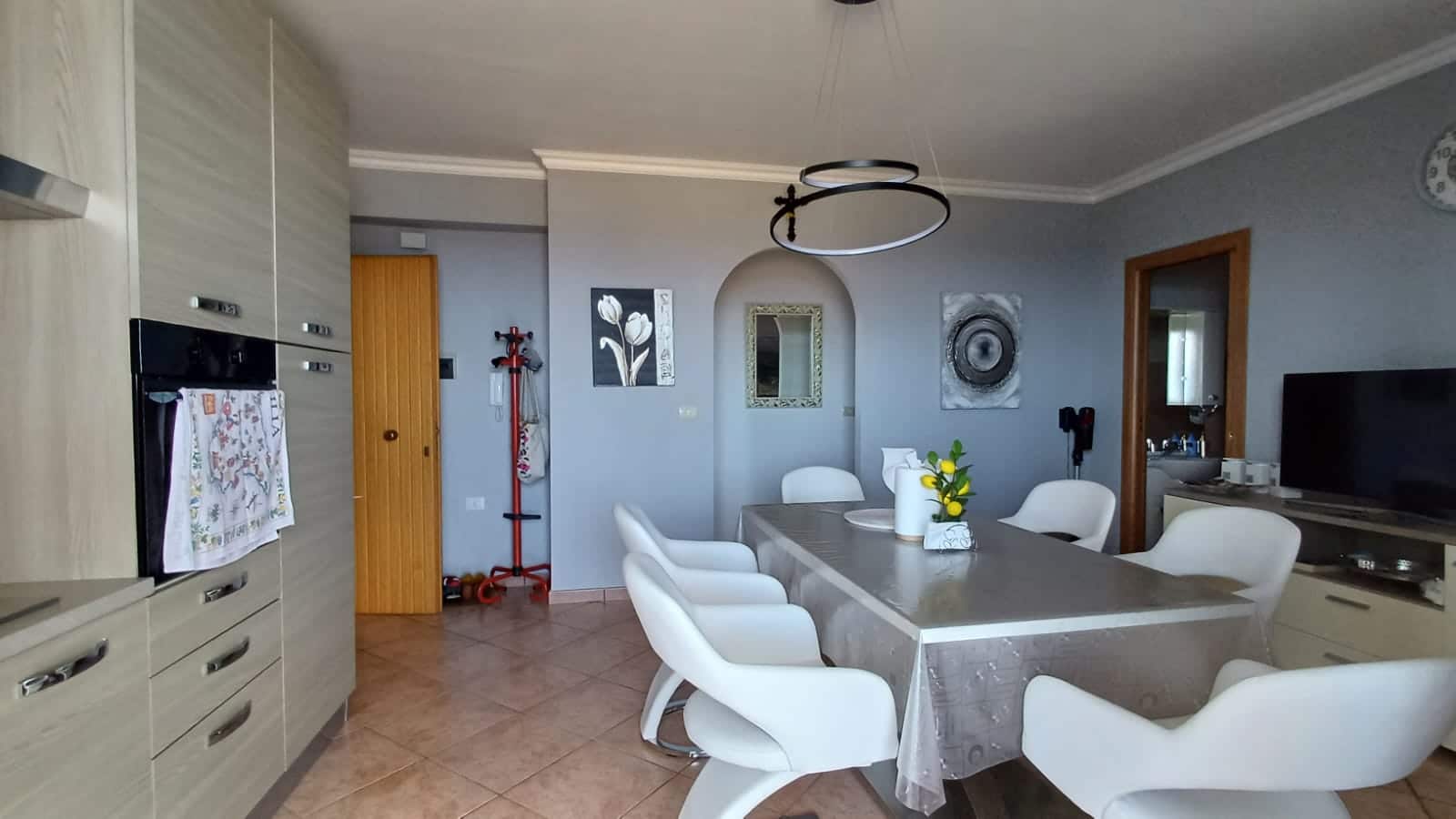 Elegante sala da pranzo con soggiorno e cucina moderna, arredi raffinati, pareti grigie, tavolo con tovaglia, sedie bianche, decorazioni artistiche e atmosfera accogliente, ideale per famiglia e amici.