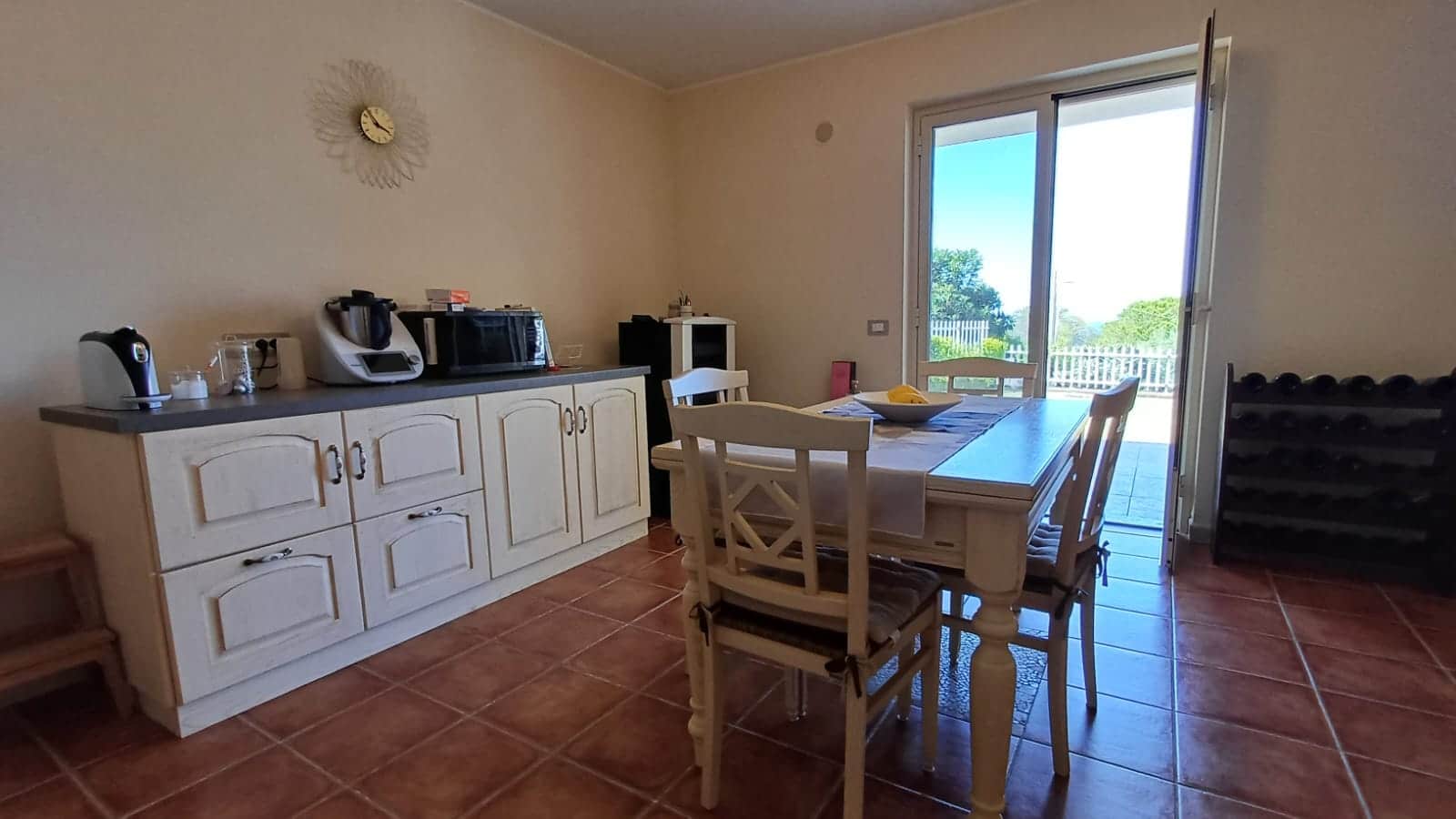Cucina abitabile con tavolo in legno, porte a vetro apribili verso un balcone con vista sul verde, mobili bianchi e cucina moderna, ambiente luminoso e accogliente.