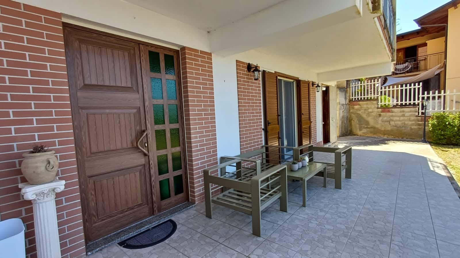 Veranda esterna con tavolo e sedie in stile moderno, accesso alla casa con grandi porte in legno e finestre con persiane in legno, ambiente accogliente e tranquillo, ideale per relax e convivialità.