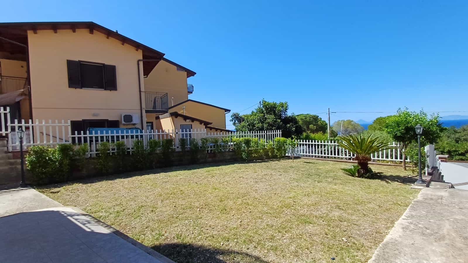 Primo piano di un giardino curato con siepi, piante ornamentali e una recinzione bianca che circonda una casa di campagna in stile rustico, con vista panoramica sulle colline e il mare in lontananza, sotto un cielo sereno e limpido.