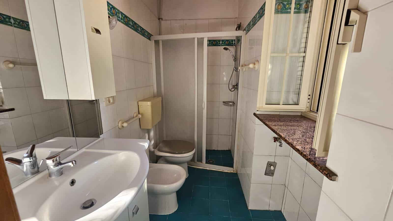 Vecchia stanza da bagno con doccia, toilette e lavabo in uno spazio compatto, finestra con tende e pareti rivestite in ceramica, atmosfera semplice e funzionale.