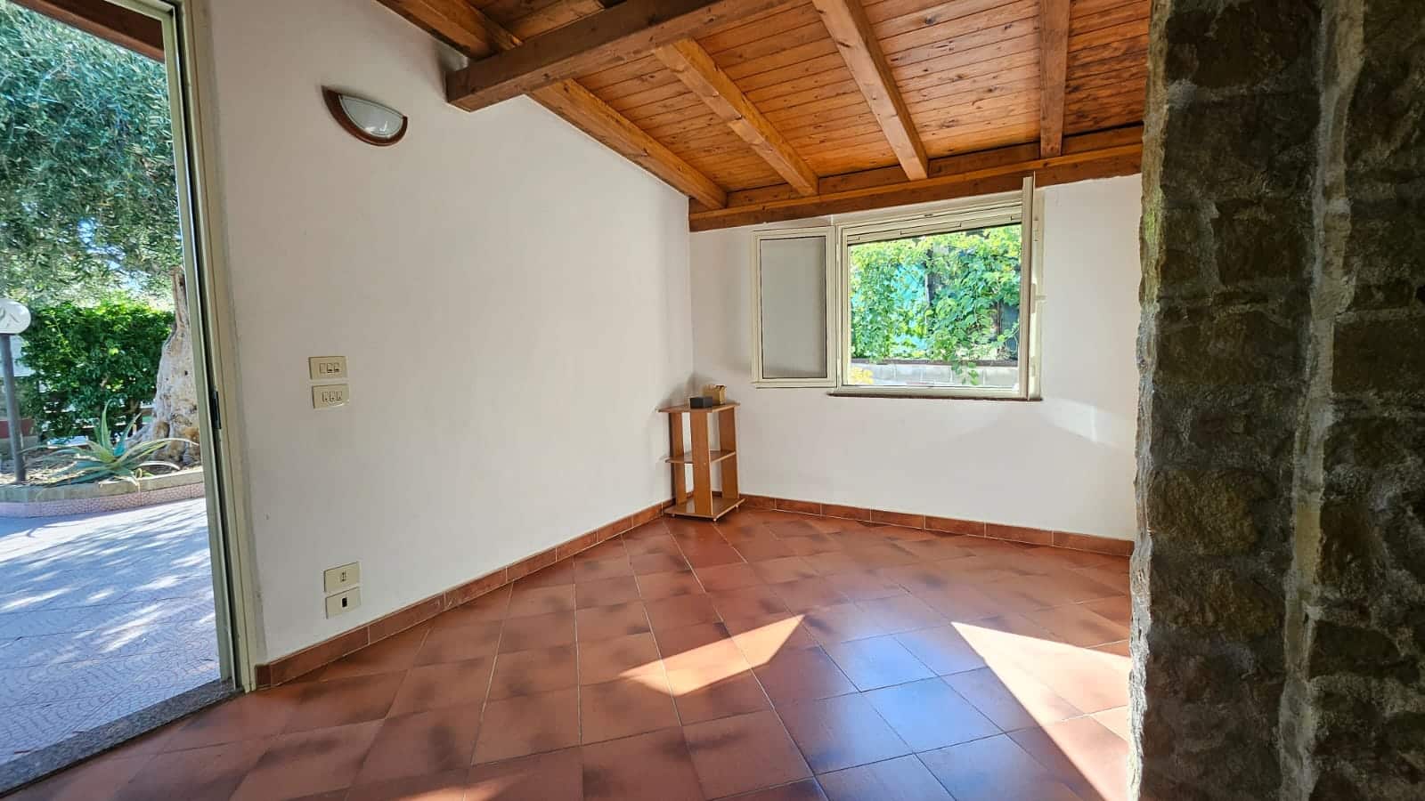 Spazio abitativo con pareti bianche, soffitto in legno e grande finestra che permette illuminazione naturale e vista sul giardino. Elementi in pietra raffinano l’ambiente, ideale per abitazioni in stile rustico o moderno.